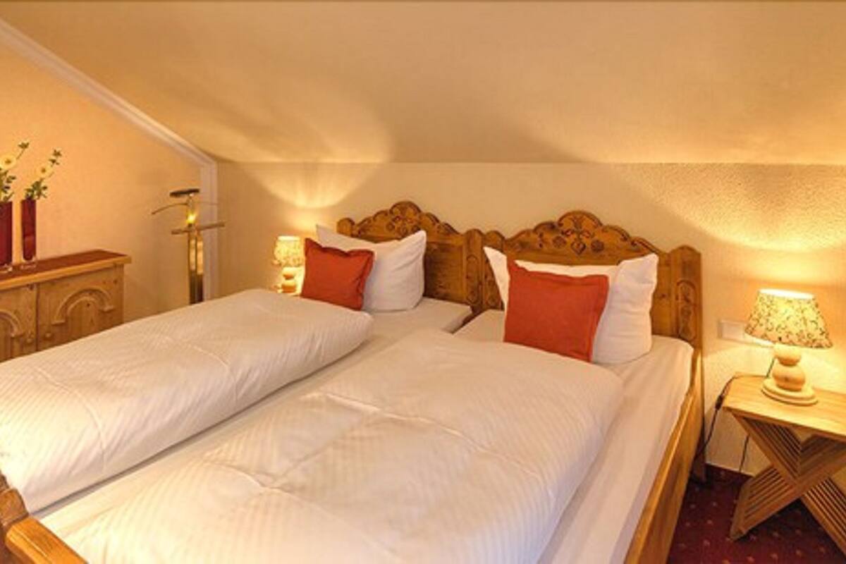 A double bed with two pillows, a wooden headboard, bedside tables with lamps, and a small wooden cabinet with plants.