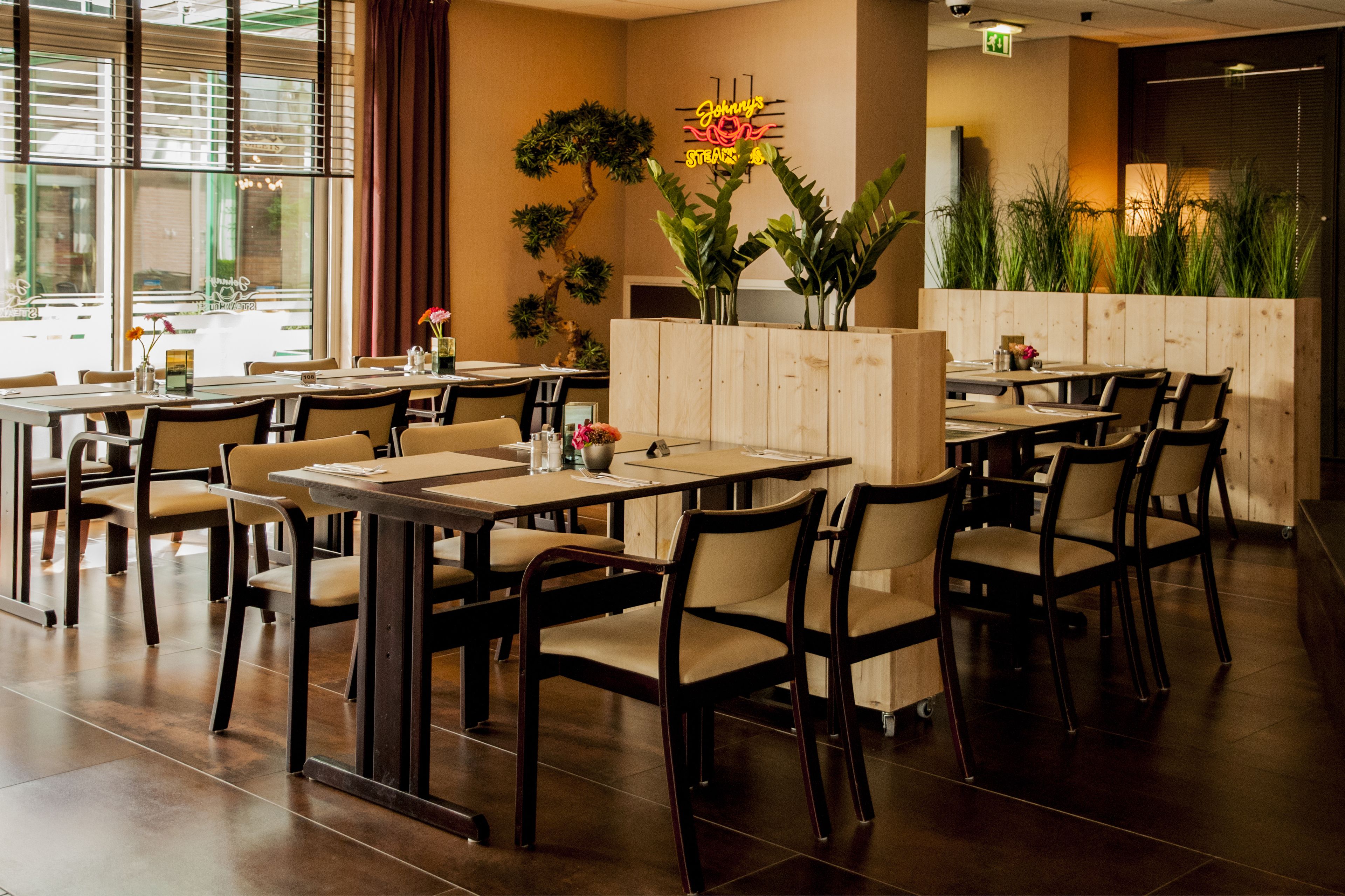 A restaurant interior with wooden tables and chairs, a neon sign, and potted plants.