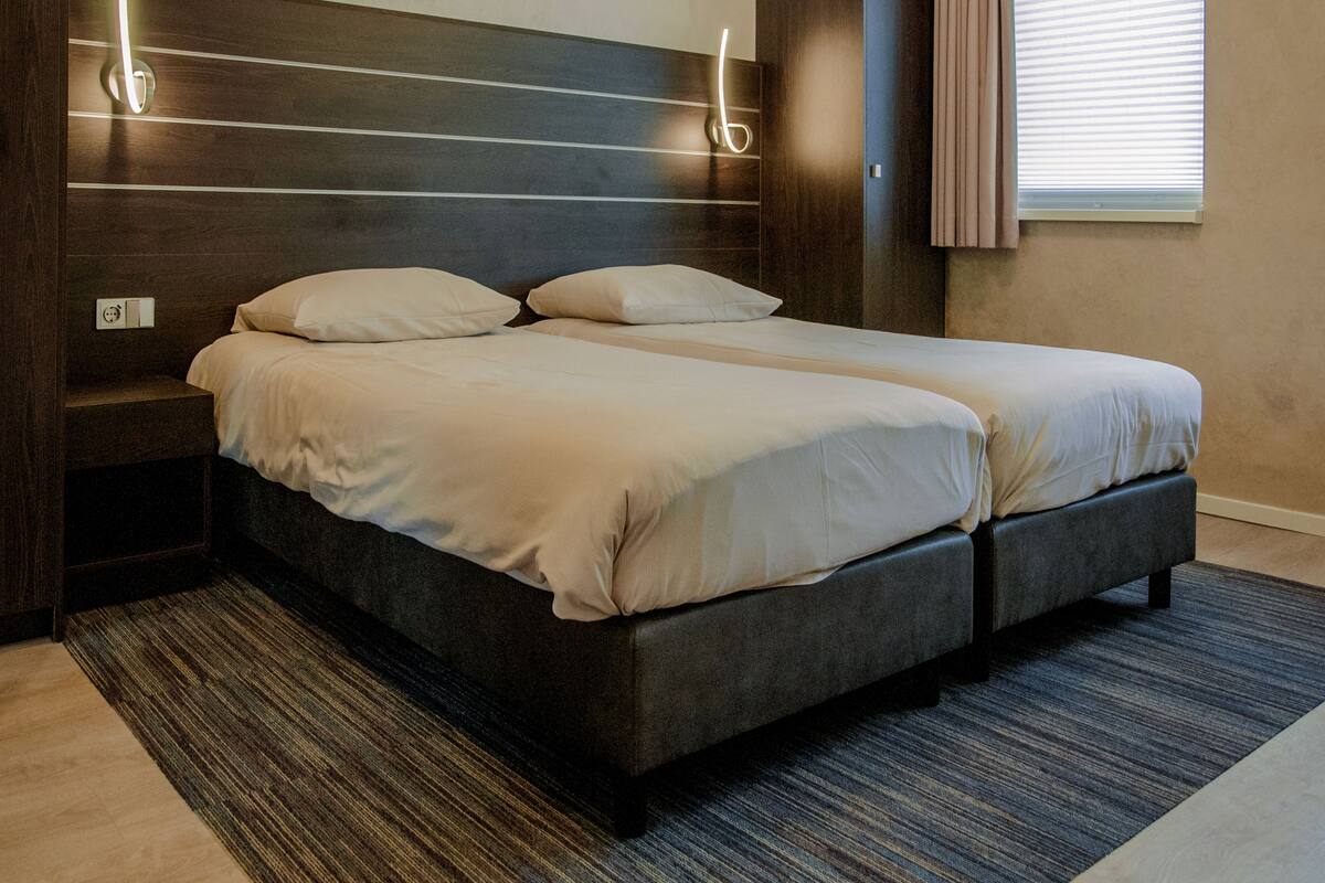 A double bed with a dark headboard and two bedside tables, a window with blinds, and a patterned rug.