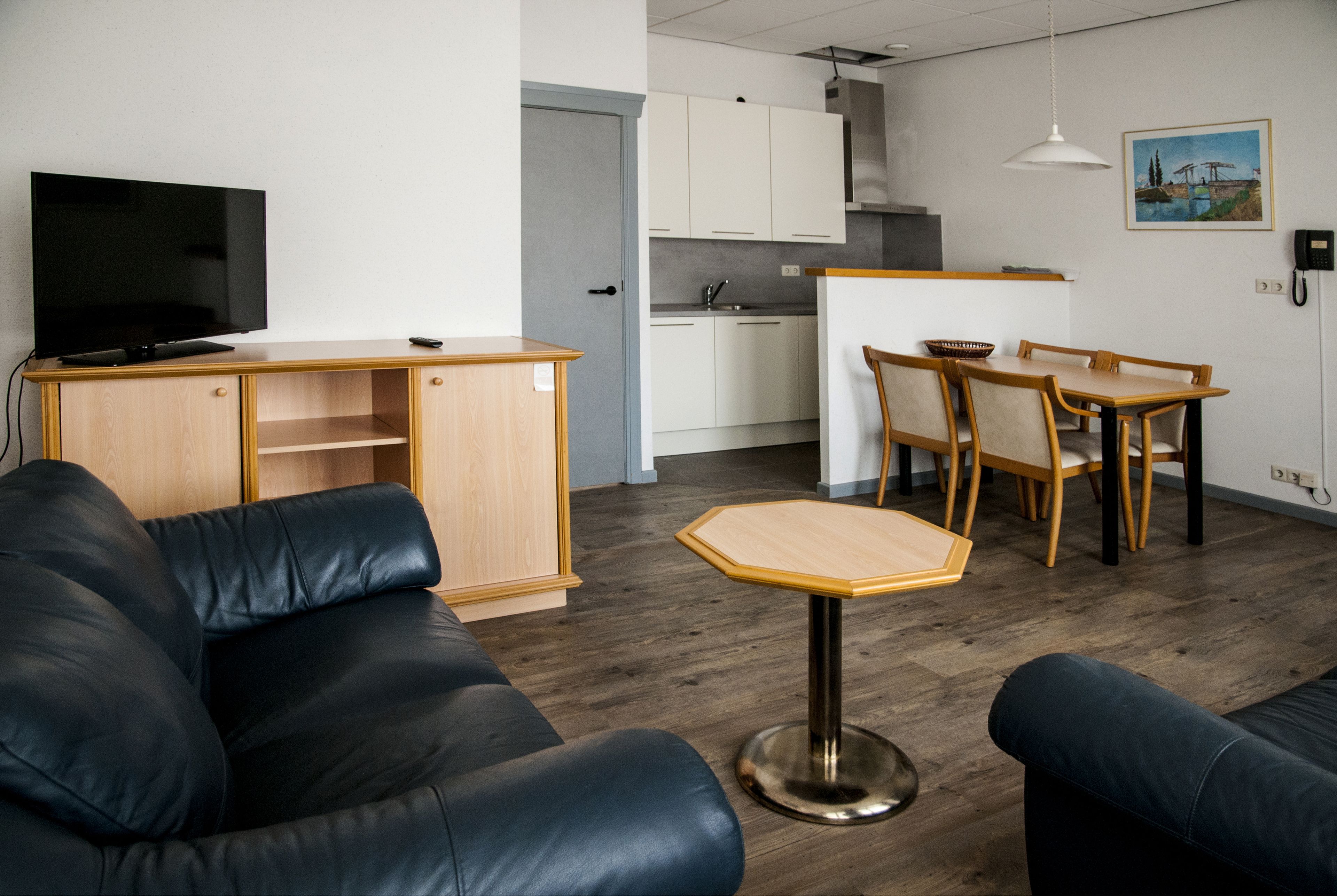 A modern living room with a black leather sofa, a wooden coffee table, and a TV on a wooden cabinet. There is a kitchen area with white cabinets and a dining table with chairs.