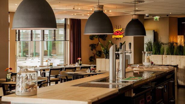 A restaurant interior with multiple dining tables, black pendant lighting, and a visible kitchen area. - Dekkers Hotel (Ossendrecht)