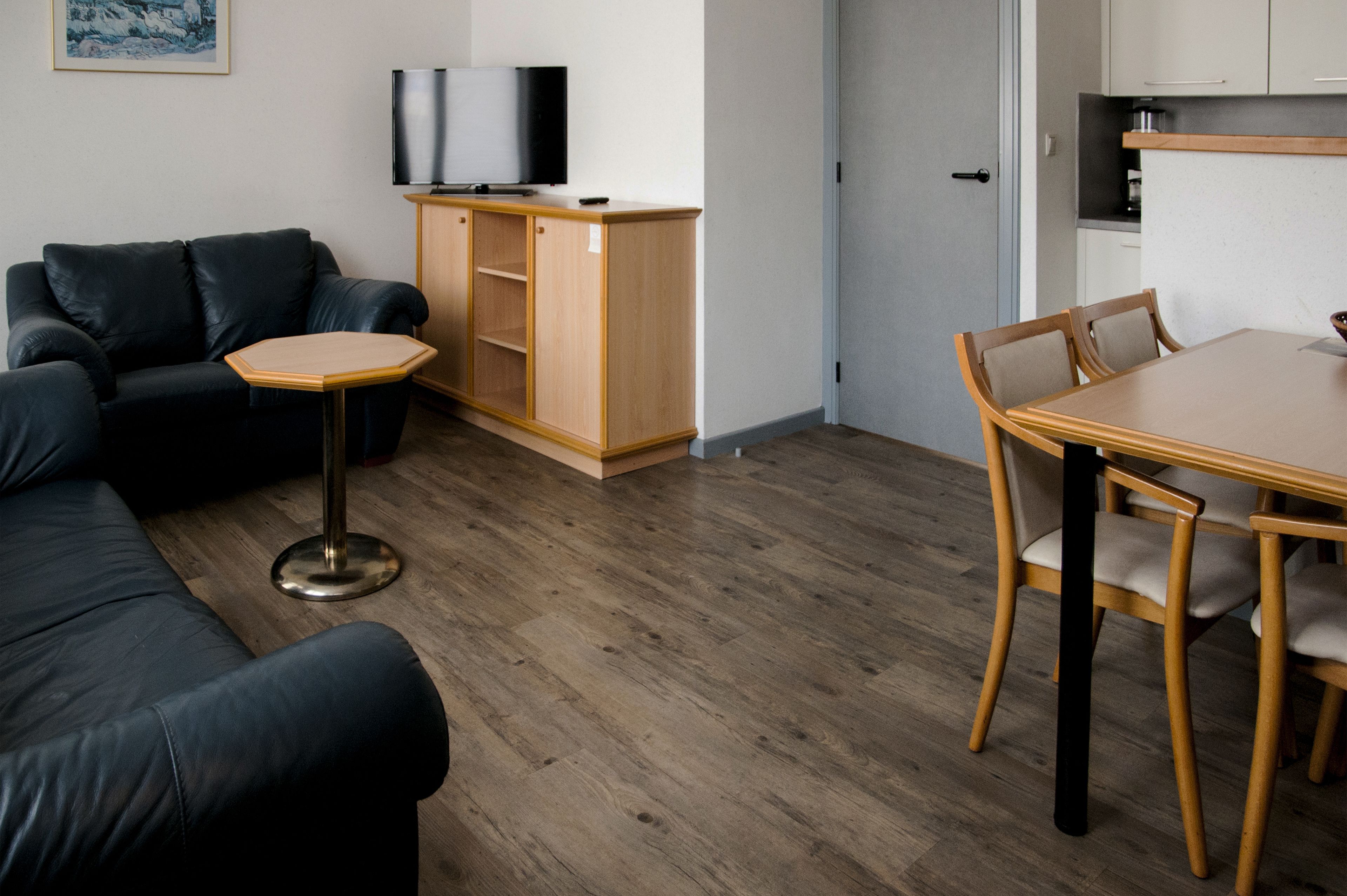 Un salon moderne avec un canapé en cuir noir, une table à manger en bois avec quatre chaises, une petite table basse ronde et un téléviseur posé sur un meuble en bois.