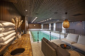 Un área de piscina interior moderna con techo de madera, una sala de estar con sofá y sillones, y un bar con mostrador de madera.