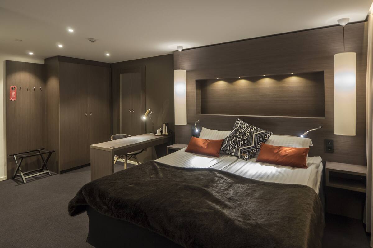 A modern hotel room with a large bed, a desk, and a chair. There are two bedside tables with lamps, a wardrobe, and a small table with a lamp.