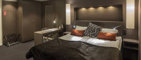 Una habitación de hotel moderna con una cama grande, un escritorio y una silla. Hay dos mesitas de noche con lámparas, un armario y una pequeña mesa con una lámpara.