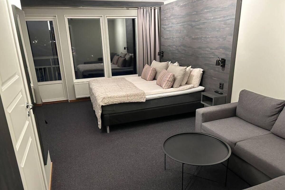 A modern hotel room with a large bed, a grey sofa, a small round table, and a view through a sliding door.