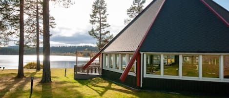 A lodge with a steep roof, large windows, and a deck overlooking a lake.
