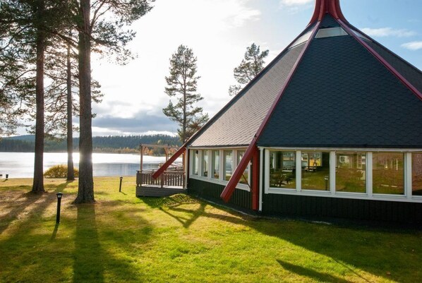 A lodge with a steep roof, large windows, and a deck overlooking a lake.