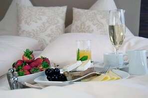 Ein liebevoll gedeckter Frühstückstisch mit Erdbeeren, Trauben, Käse und einem Glas Saft auf einem Bett mit weißen Leinentüchern und gemusterten Kissen.