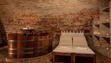 Ein Zimmer mit Steinwänden, einem hölzernen Bett und einem fassförmigen Gegenstand, vermutlich einem Weinfass, sowie einem kleinen Tisch und Kerzen.