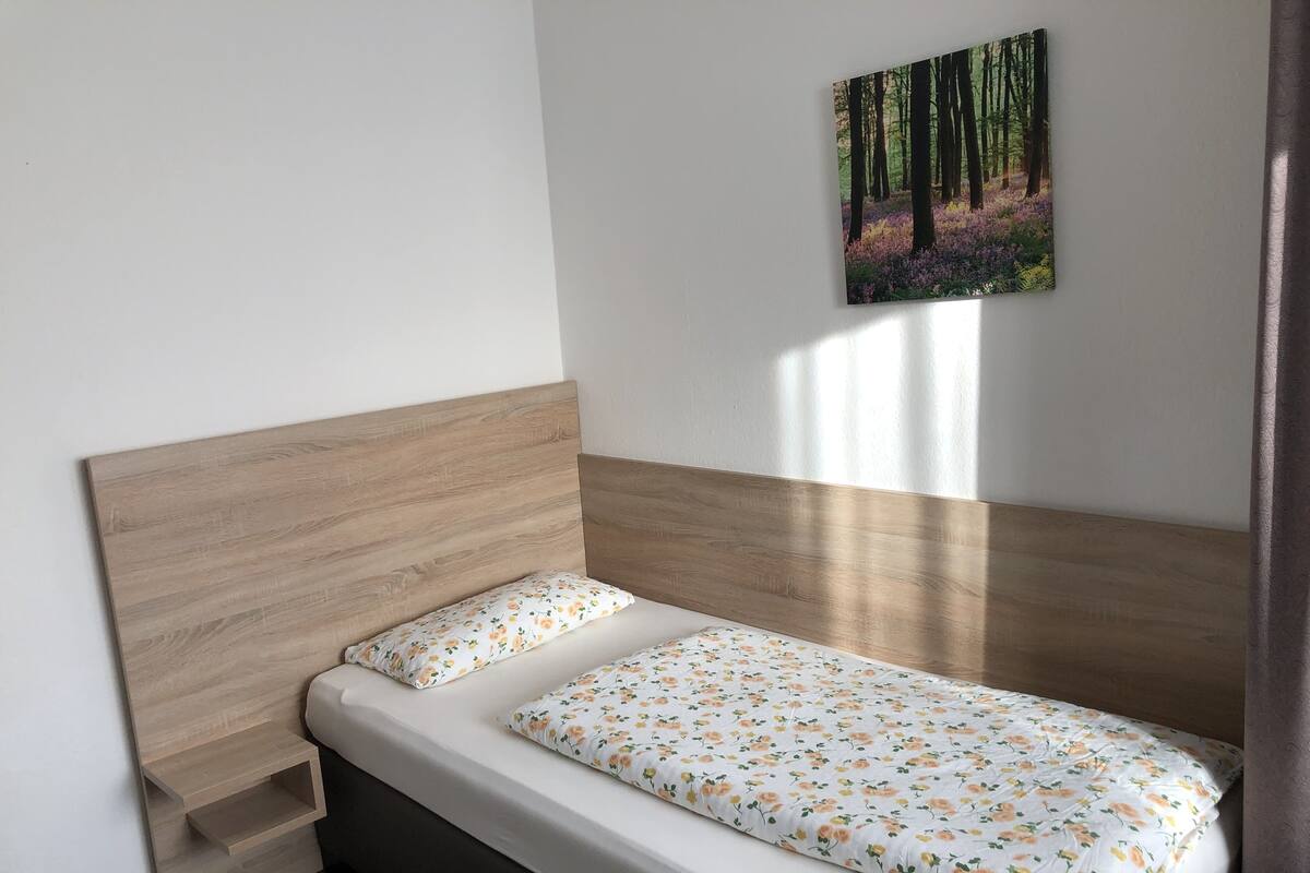 A single bed with a wooden headboard, white bedding with a floral pattern, and a painting of a forest on the wall.