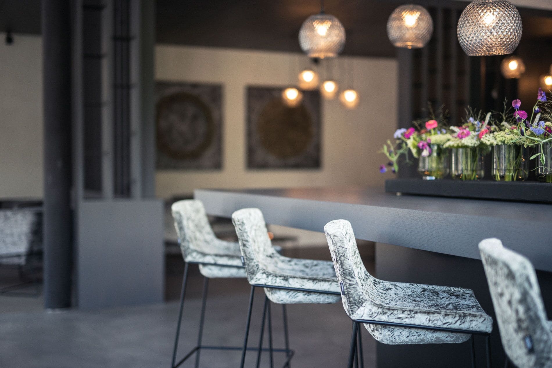 Een moderne bar met drie witte barstoelen, een donkere bar en decoratieve hanglampen.