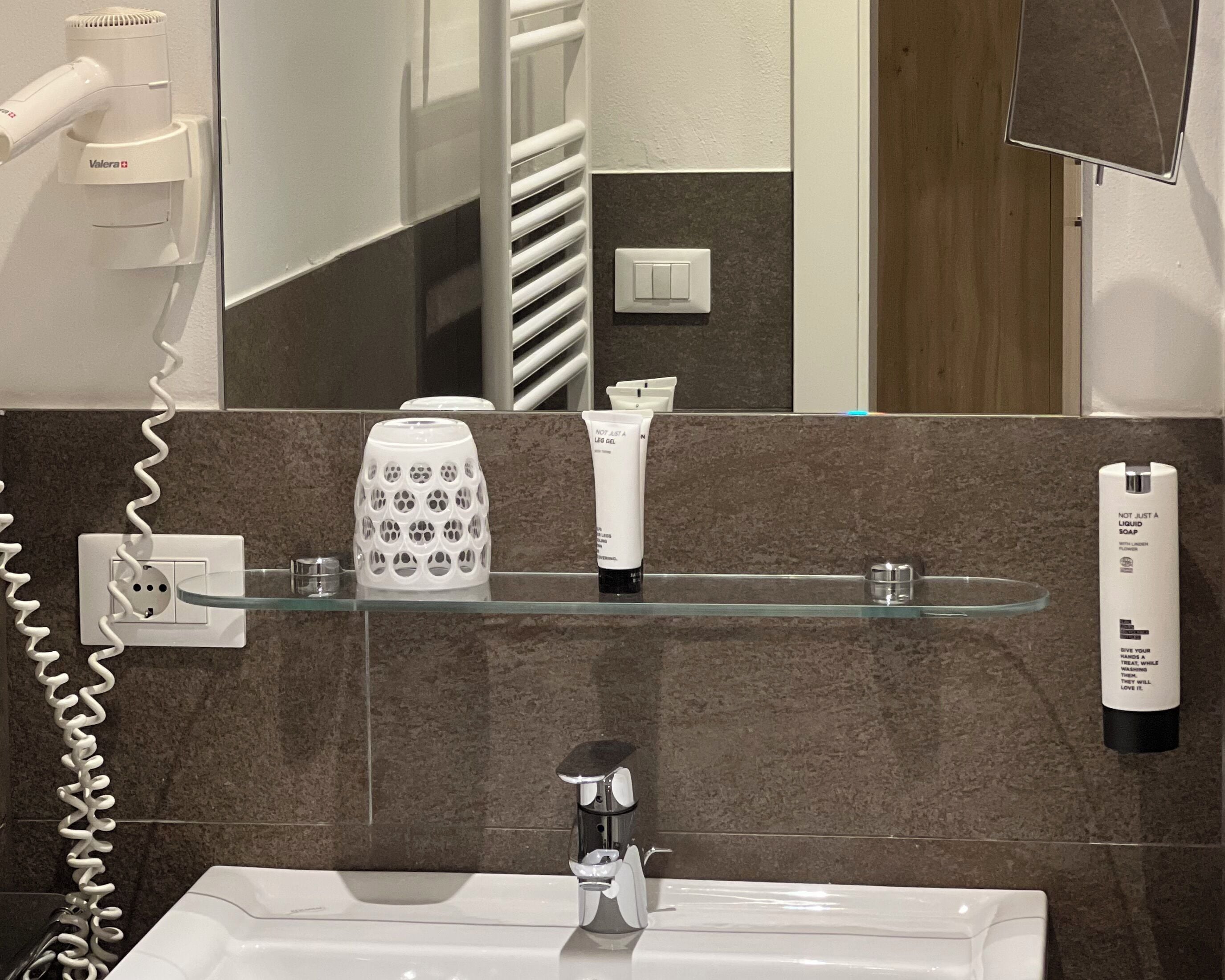A bathroom with a white sink, a mirror, a towel rack, a glass soap dispenser, and a hairdryer on the wall.