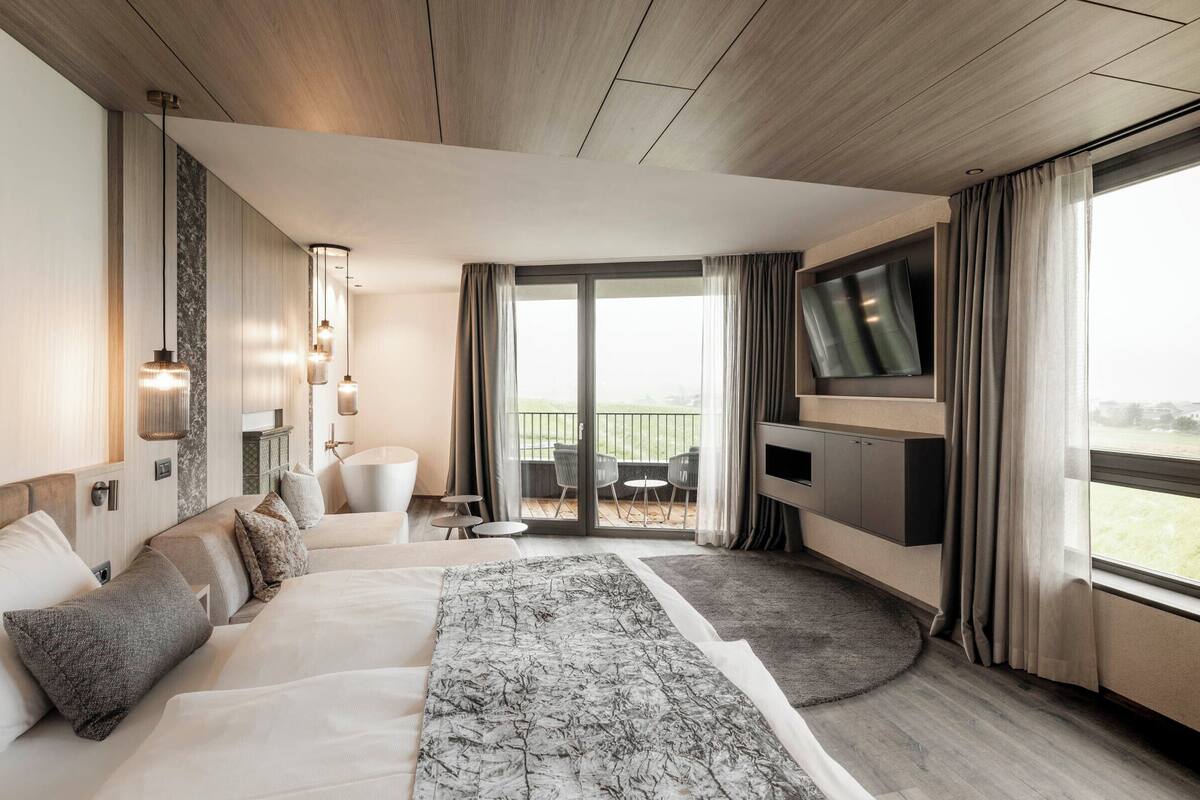 A modern hotel room with a large bed, a TV, a balcony with outdoor seating, and a view of the outdoors.