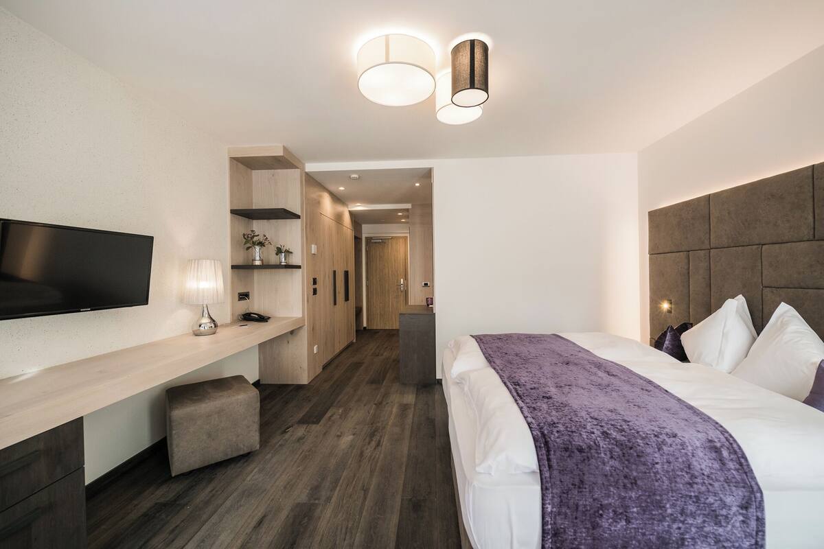 A modern hotel room with a flat-screen TV, a bedside table with a lamp, a bed with a headboard, and a hallway leading to another room.