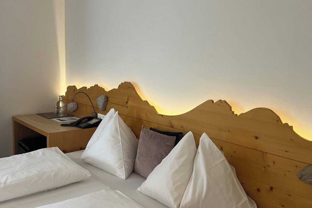 A neatly made bed with white linens and pillows, a wooden headboard, a bedside table with a lamp, and a nightstand with a phone and a bottle.