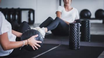 Una persona sentada sobre una esterilla, sosteniendo una pelota medicinal, con equipo de gimnasio y kettelbells al fondo.