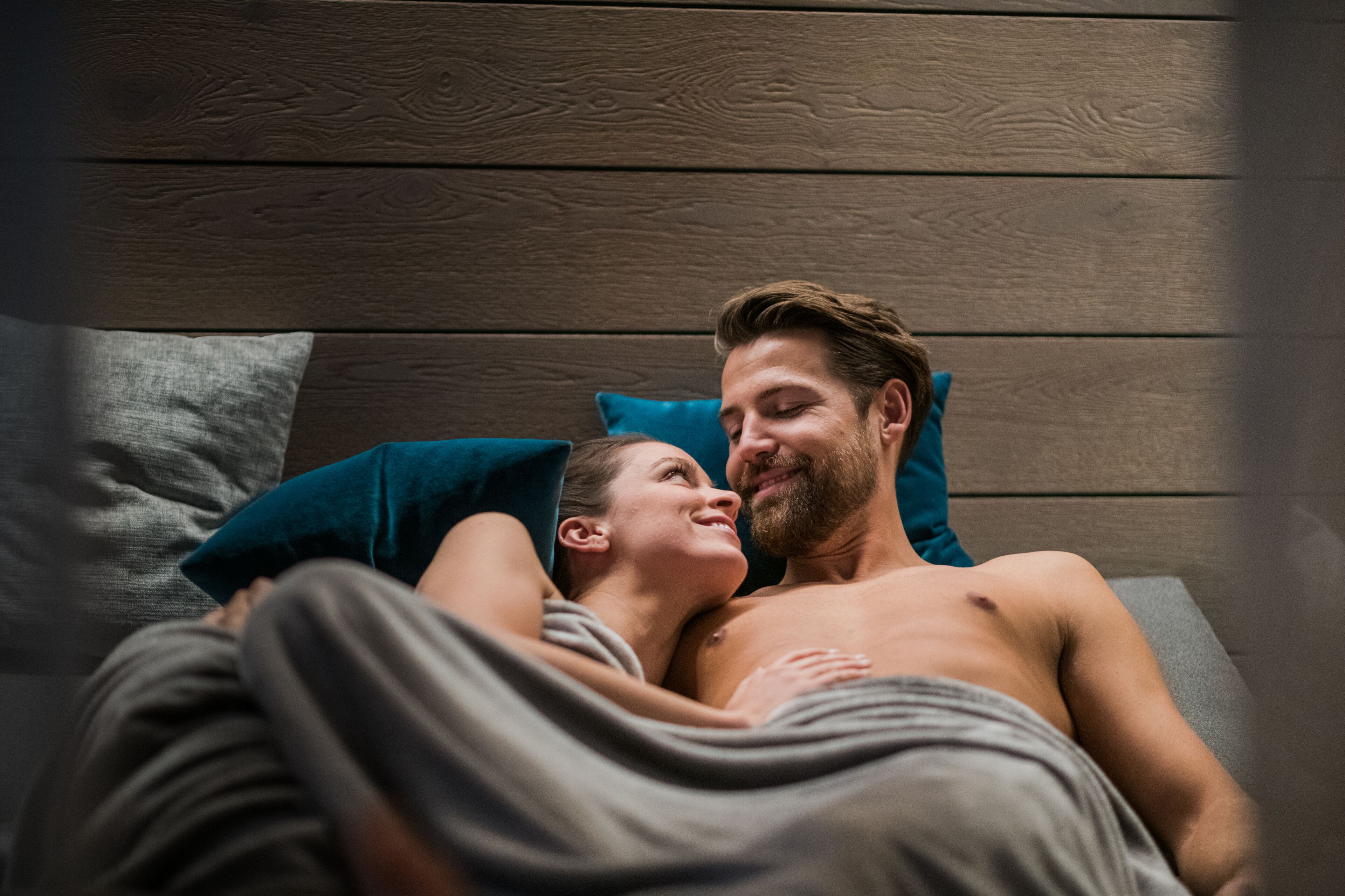 Een koppel ligt op een bed met een houten hoofdbord, bedekt met een grijze deken.