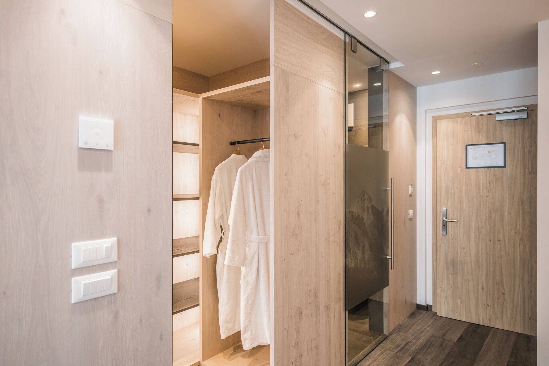 Un baño moderno con paredes de madera, cabina de ducha con mampara de vidrio y una bata blanca colgada en un perchero.