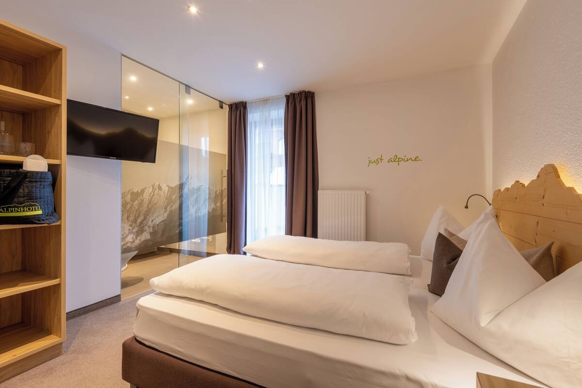 A modern hotel room with a large bed, a TV, a wooden shelf with items, and a mountain view.