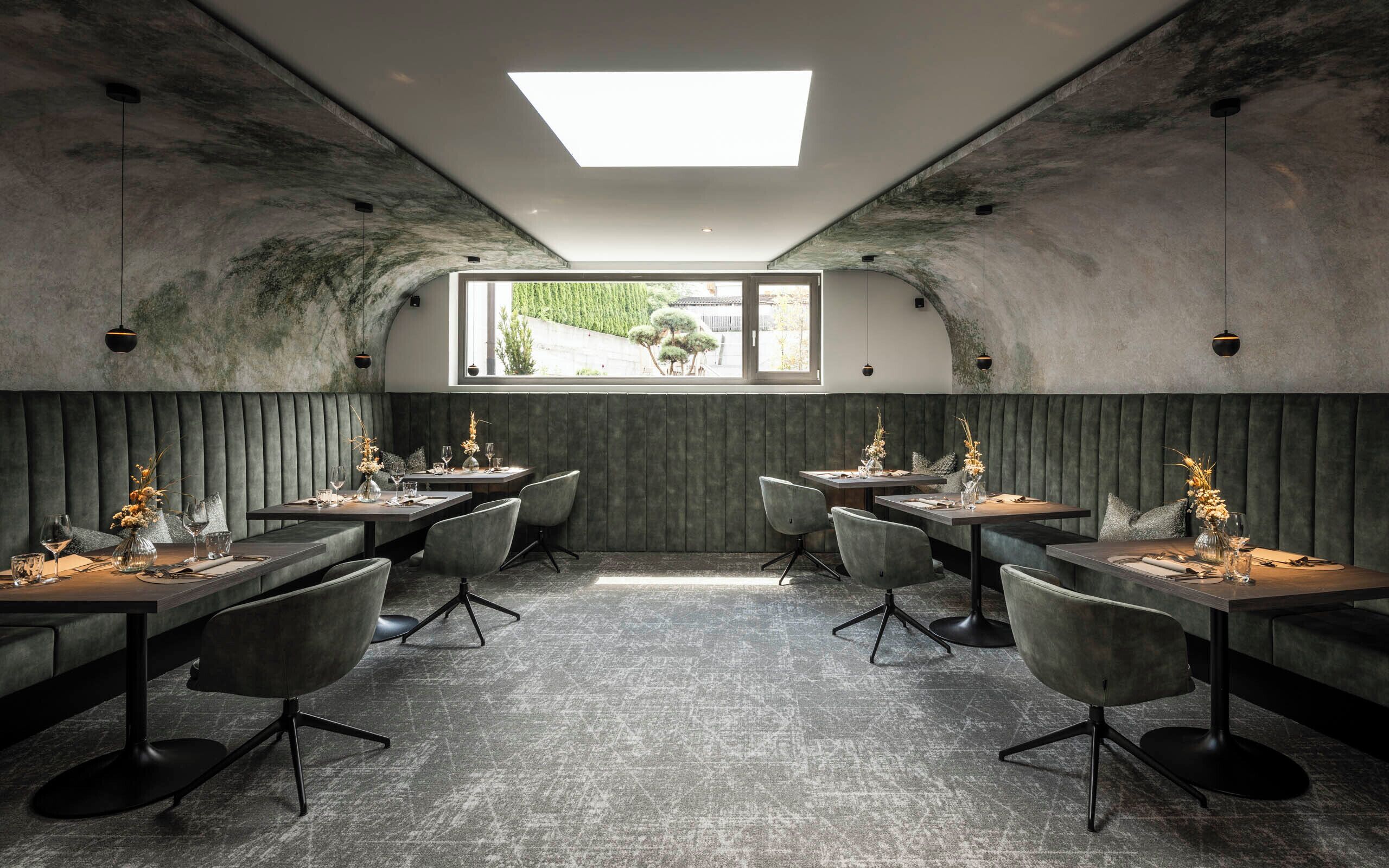 Ein Restaurant mit modernem Ambiente, das einen zentralen Essbereich mit Tischen und Stühlen, ein großes Fenster mit Blick ins Freie sowie ein außergewöhnliches Decken-Design mit Oberlicht bietet.