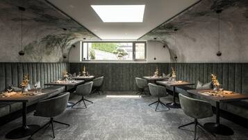 Ein Restaurant mit modernem Ambiente, das einen zentralen Essbereich mit Tischen und Stühlen, ein großes Fenster mit Blick ins Freie sowie ein außergewöhnliches Decken-Design mit Oberlicht bietet.