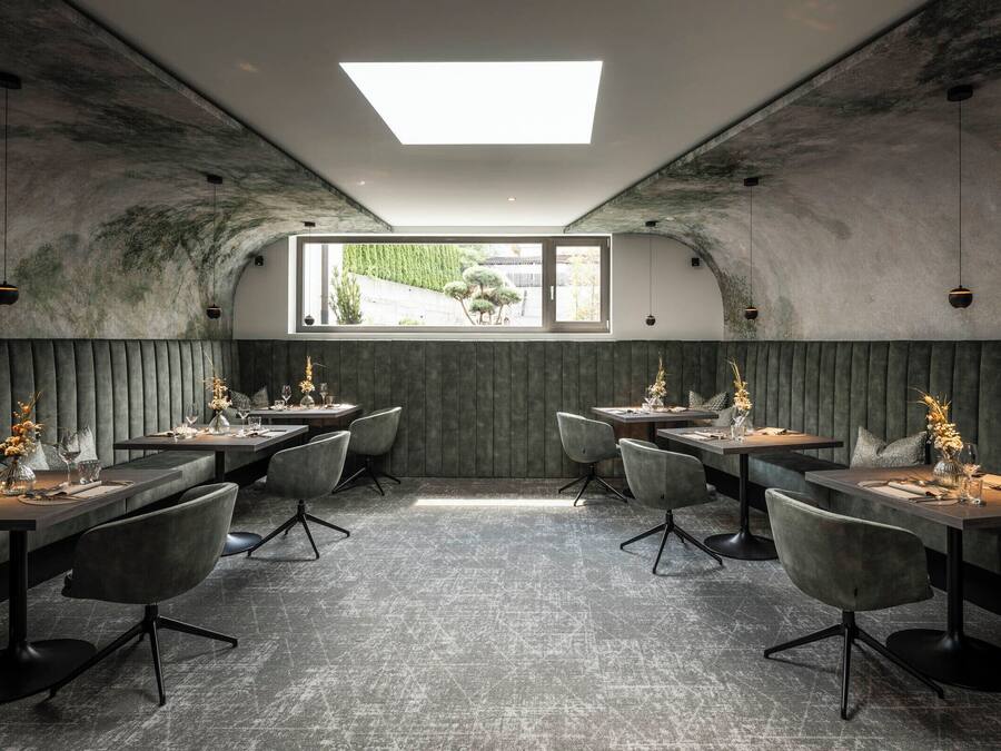 Ein Restaurant mit modernem Ambiente, das einen zentralen Essbereich mit Tischen und Stühlen, ein großes Fenster mit Blick ins Freie sowie ein außergewöhnliches Decken-Design mit Oberlicht bietet.