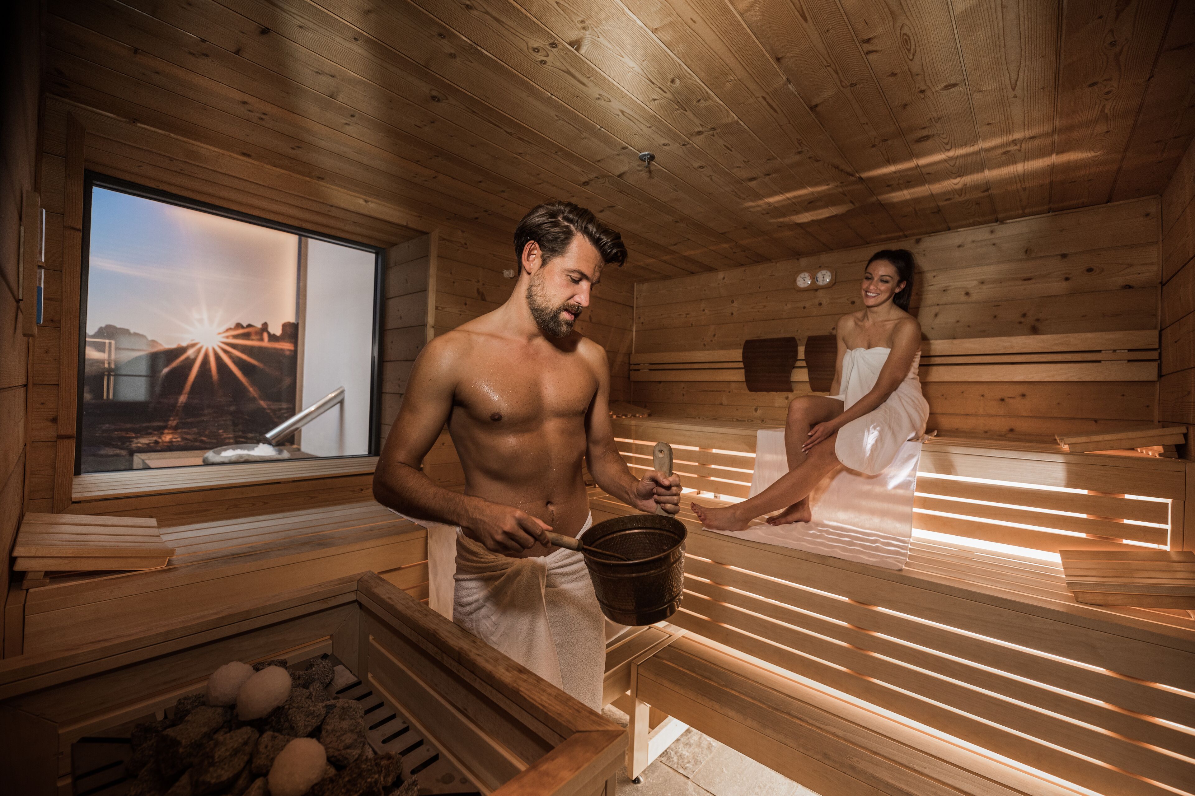 Eine Sauna mit Holzwänden und -bänken, ein Fenster mit Sonnenuntergangblick und ein Eimer mit Steinen.