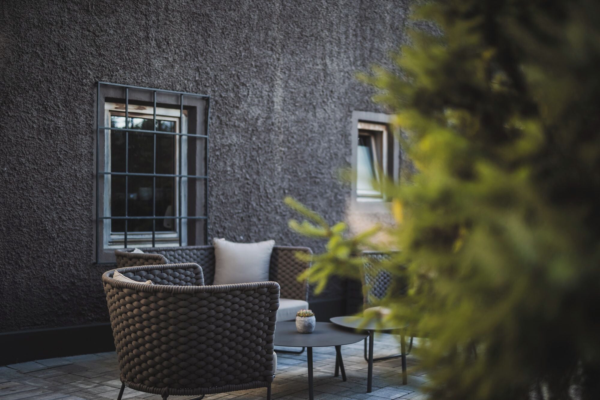 Un patio con mobiliario de mimbre y una pequeña mesa, frente a una pared texturizada con ventanas.