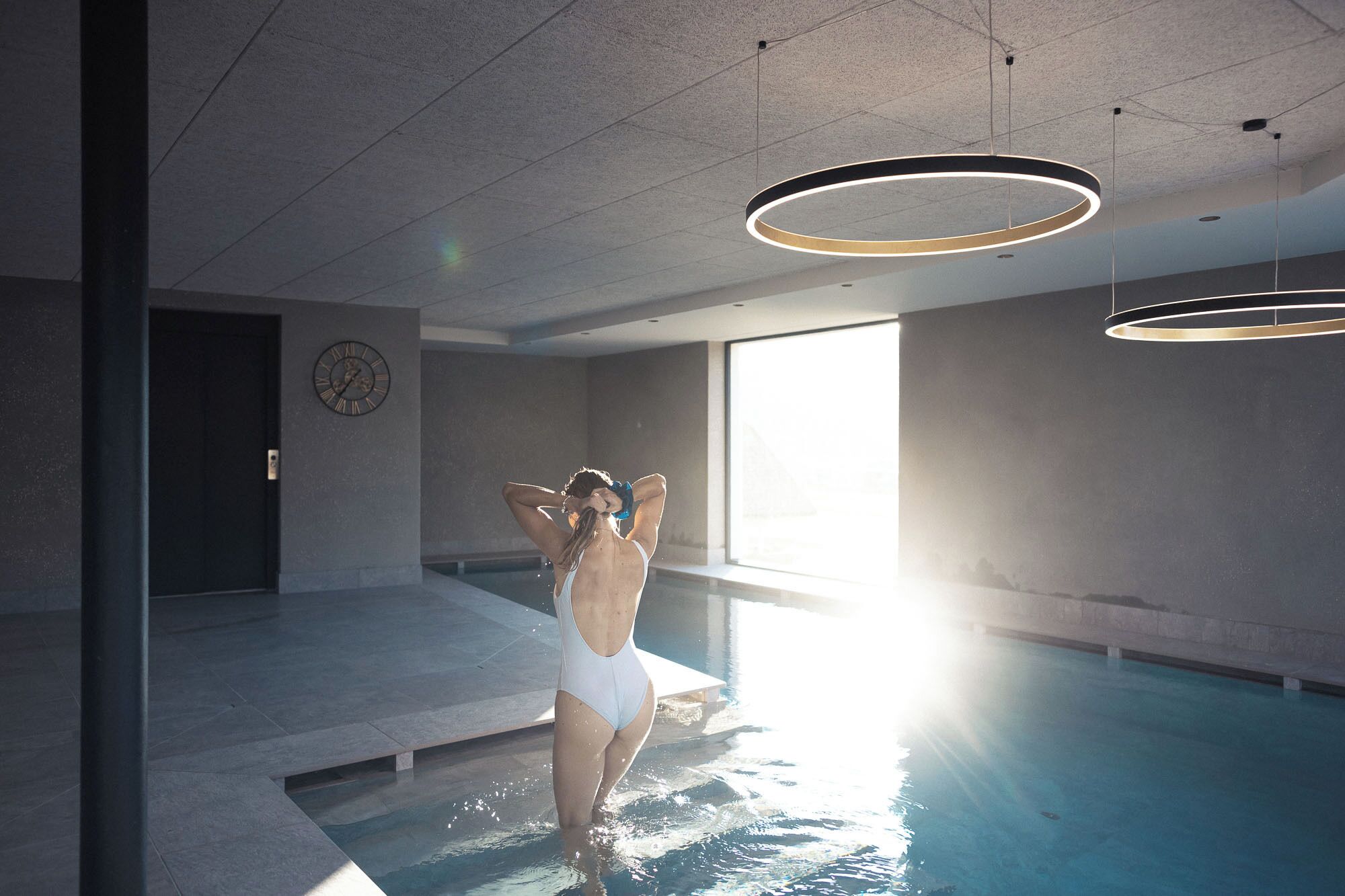 A modern indoor swimming pool with a minimalist design, featuring a large window and a visible clock on the wall.