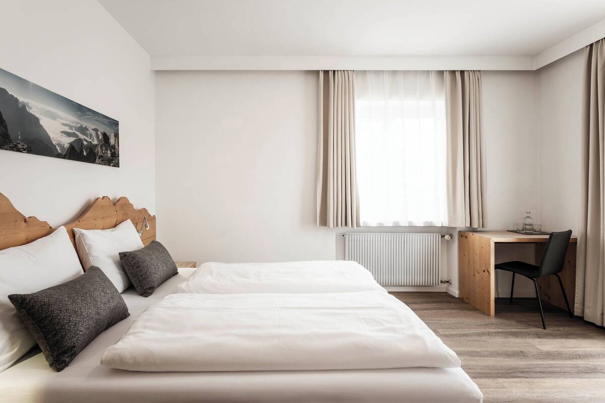 A hotel room with a bed, a wooden headboard, a desk, a chair, a radiator, and a window with curtains.