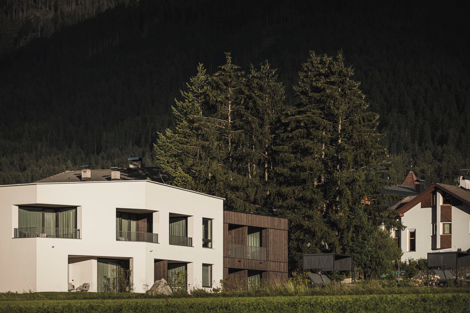 Un edificio moderno con varios balcones y ventanas amplias, rodeado de un bosque denso y montañas.