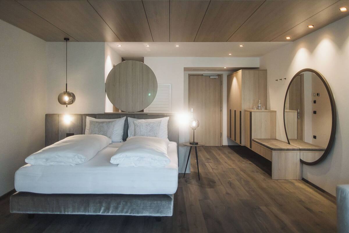 A modern bedroom with a large bed, bedside lamps, a round mirror, and a wooden wardrobe.