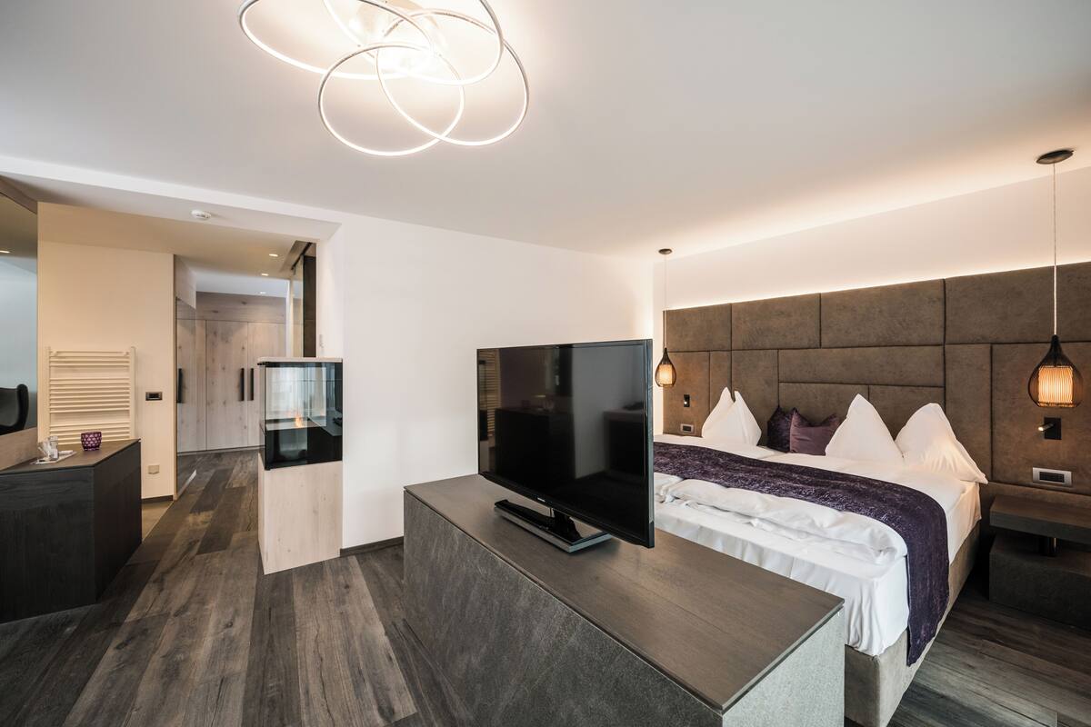 A modern hotel room with a flat-screen TV, a bed with white linens, a wooden headboard, and a built-in desk with a flat-screen TV.