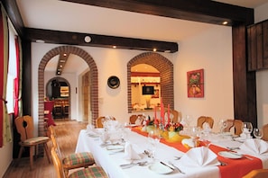 Une salle à manger avec une longue table dressée pour un repas, des chaises en bois et un mur en briques agrémenté d’une arche.