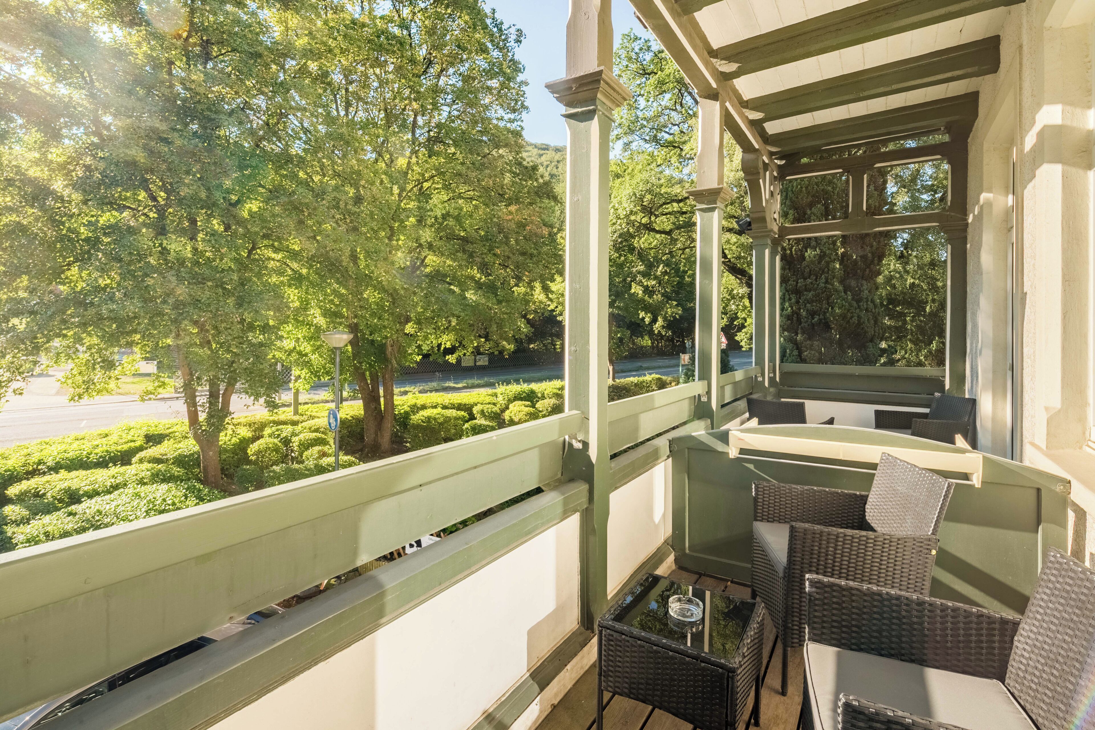 Un balcon agrémenté de meubles en osier, d’une table en verre et offrant une vue sur un jardin bien entretenu et sur la rue.