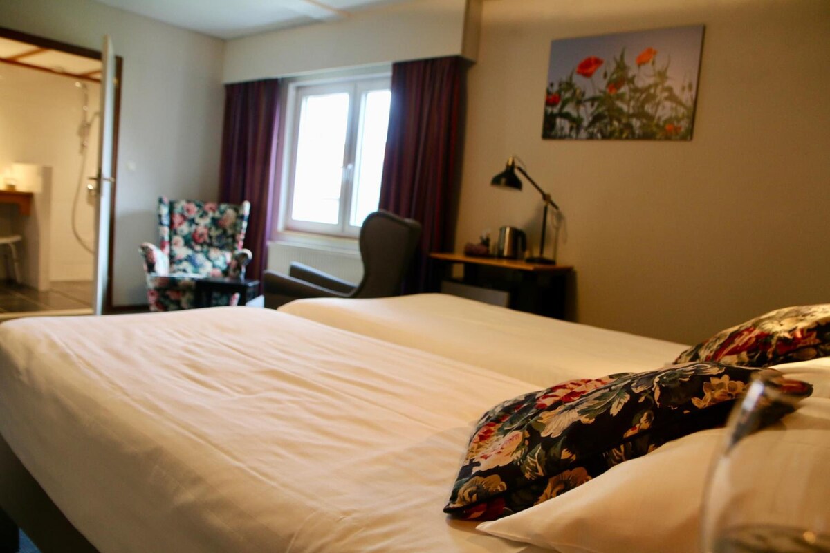 A hotel room with a bed, a desk, a chair, a window with curtains, and a painting on the wall.