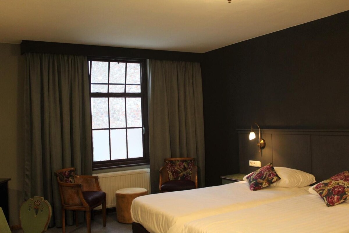 Een hotelkamer met een bed, twee stoelen, een klein tafeltje, een raam met gordijnen en een wandlamp.