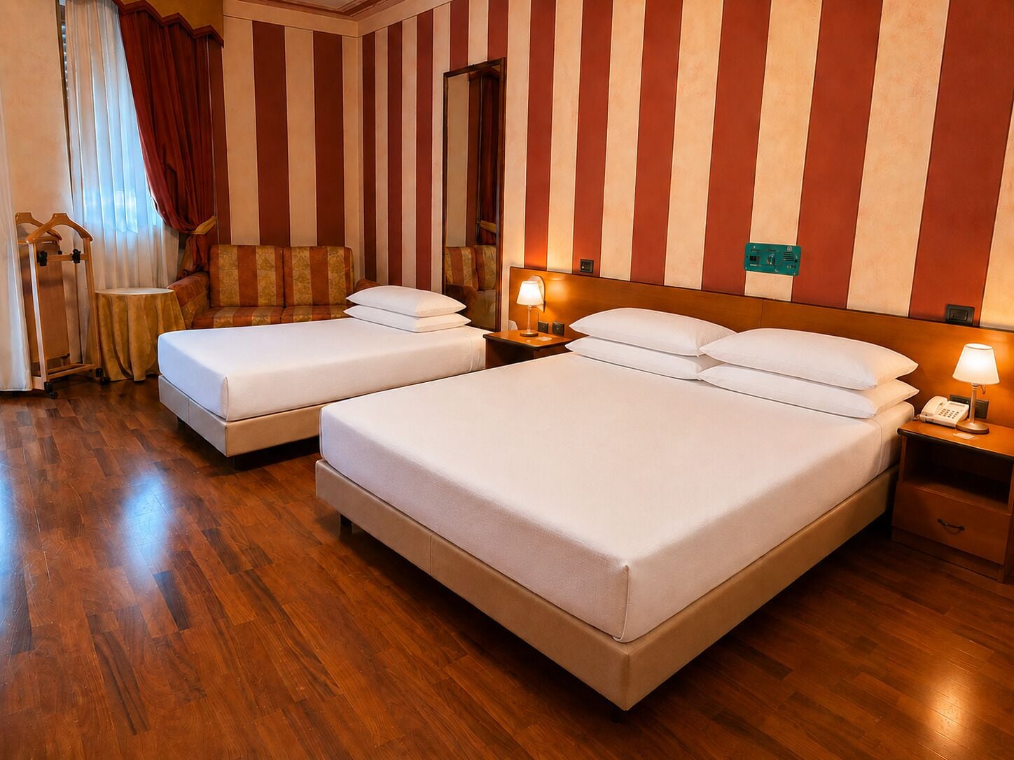 A hotel room with two beds, wooden flooring, striped walls, a mirror, and a bedside table with a lamp.