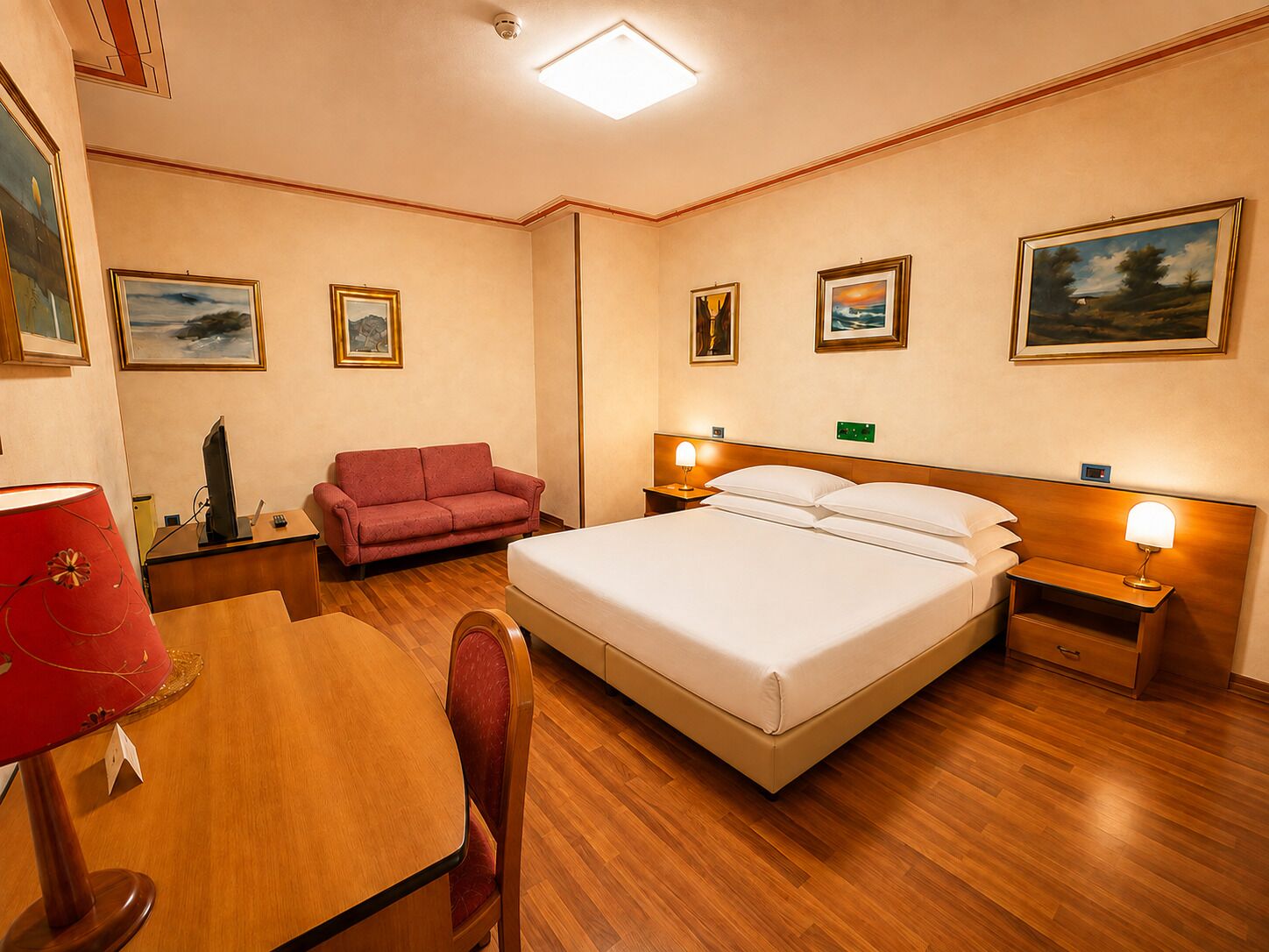 A hotel room with a bed, a desk, a chair, a red sofa, and artwork on the walls.