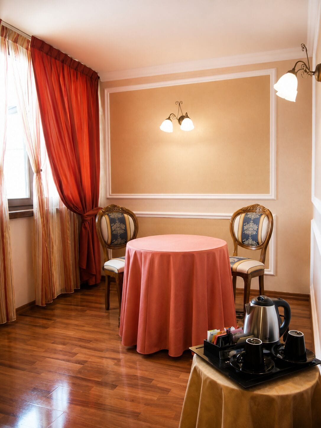 Une pièce avec une table ronde recouverte d’une nappe rose, deux chaises à l’assise à motifs, un luminaire fixé au mur et une petite table sur laquelle se trouvent une bouilloire et des tasses.
