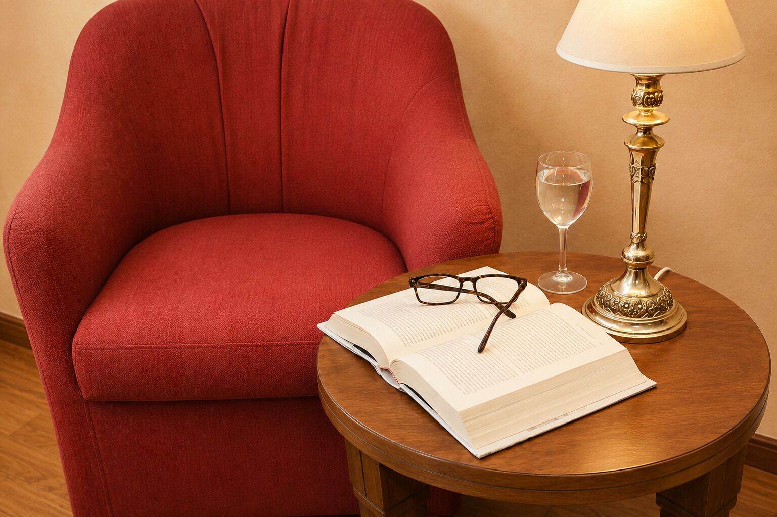 Un sillón rojo, una mesa de madera redonda con un libro, gafas, una copa de vino y una lámpara.