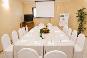 Un salón de conferencias con una larga mesa, sillas, una pantalla de proyector, un televisor y una planta.