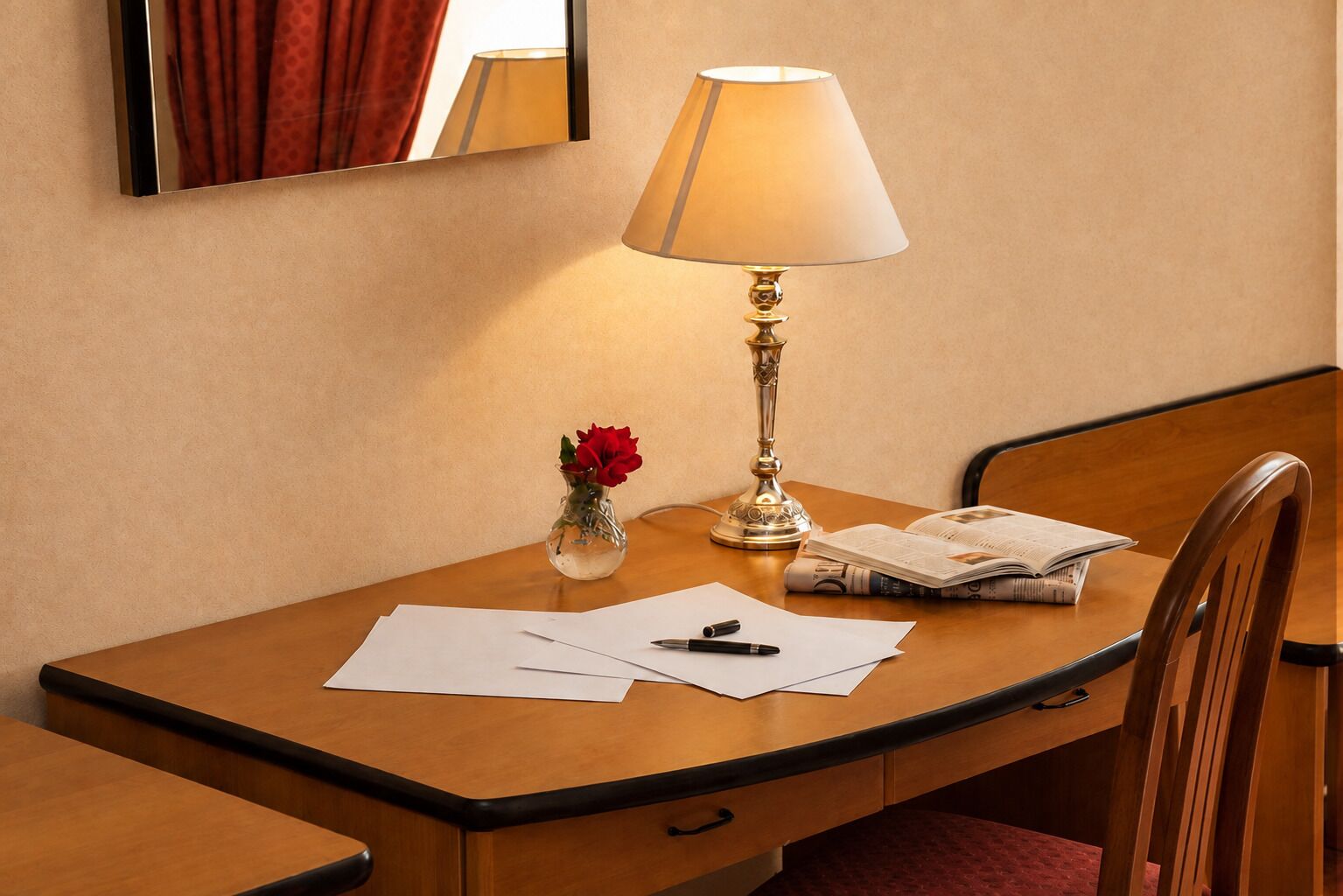 Ein Schreibtisch im Hotelzimmer mit einer Lampe, einer Vase mit einer roten Blume, Papieren, einem Stift und einem Buch.