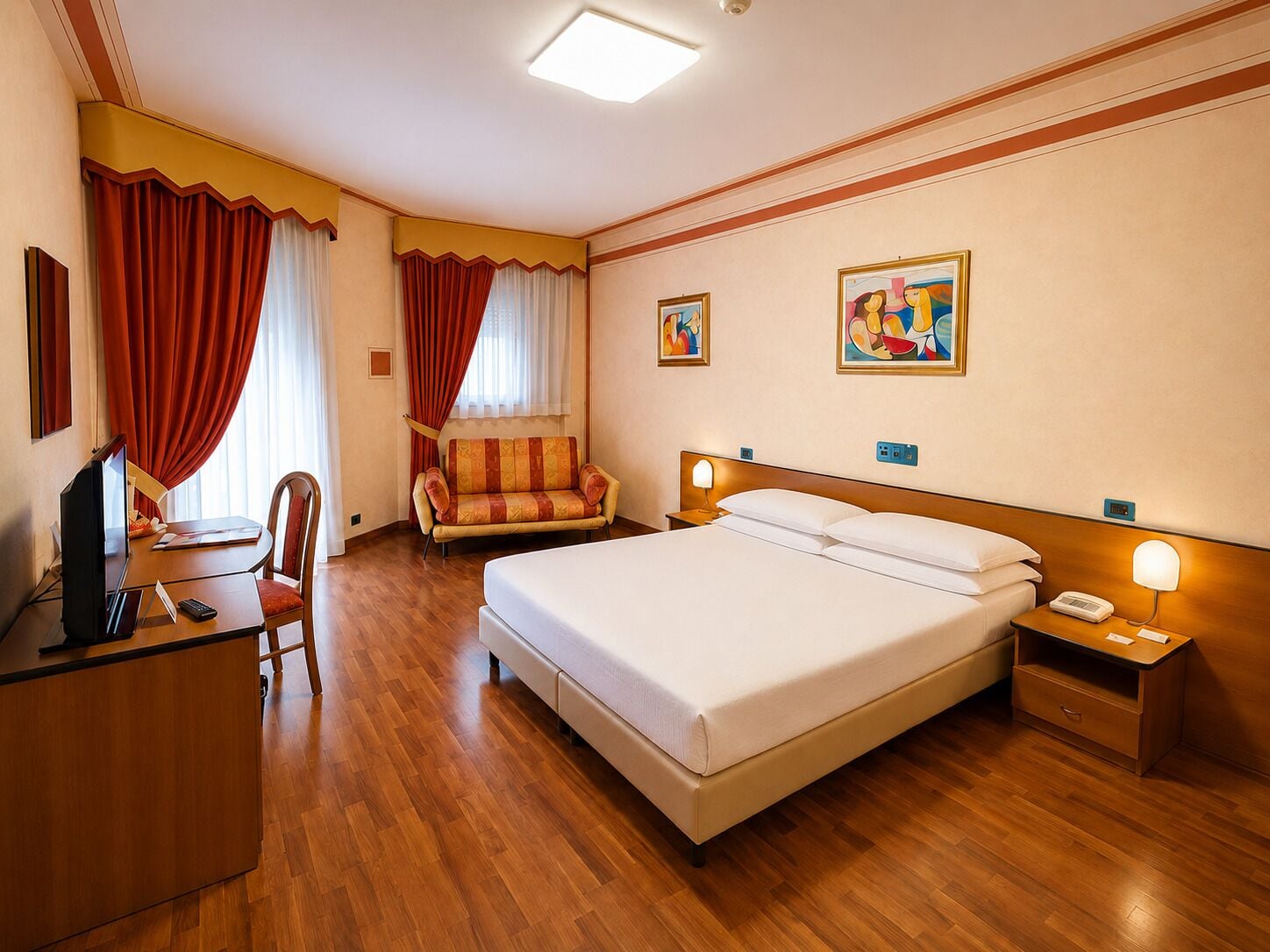 Quarto de hotel com cama, escrivaninha com computador, poltrona, sofá e duas pinturas na parede.
