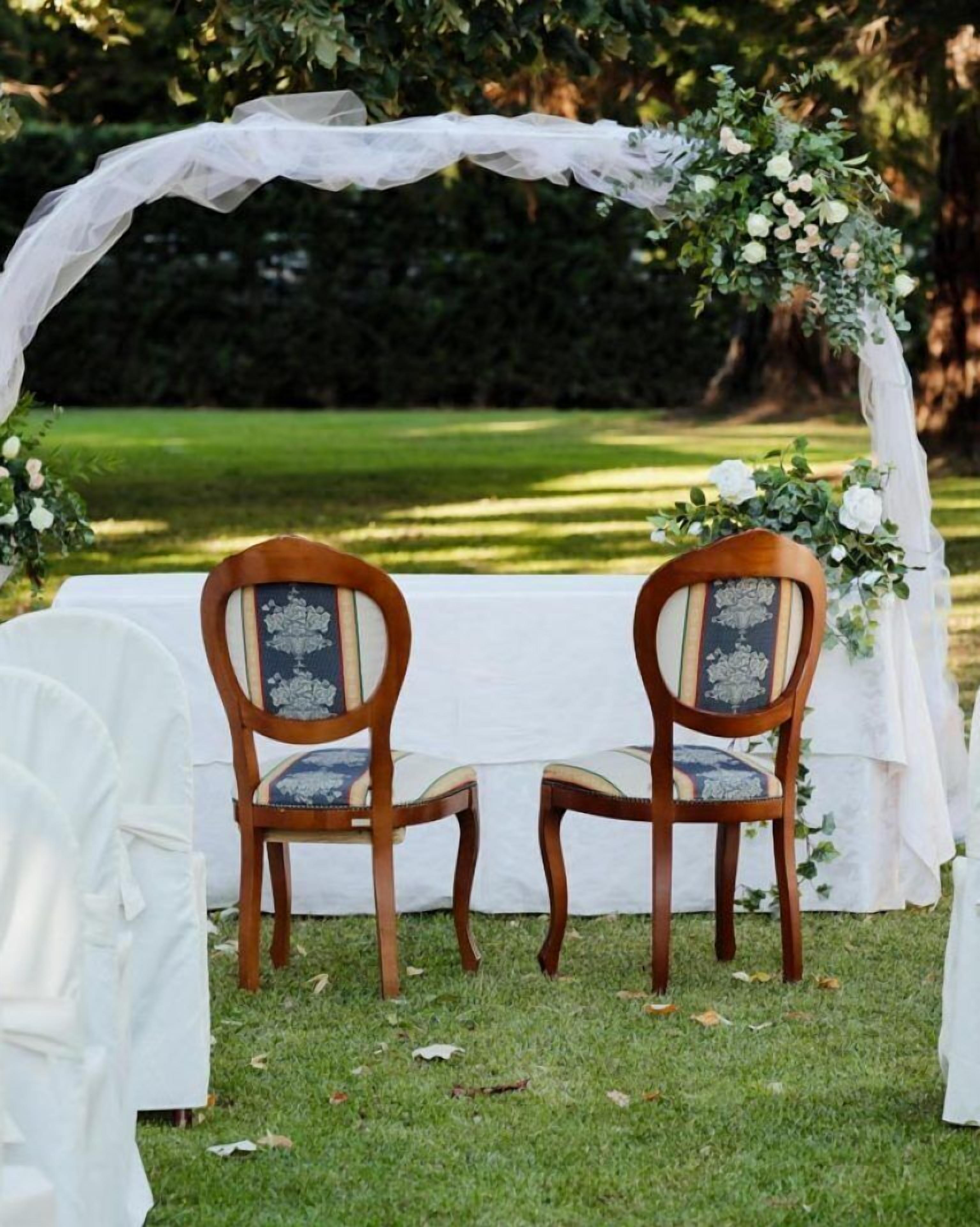 Ein Hochzeitbogen mit floralen Dekorationen und zwei hölzerne Stühle mit gepolsterten Sitzflächen.