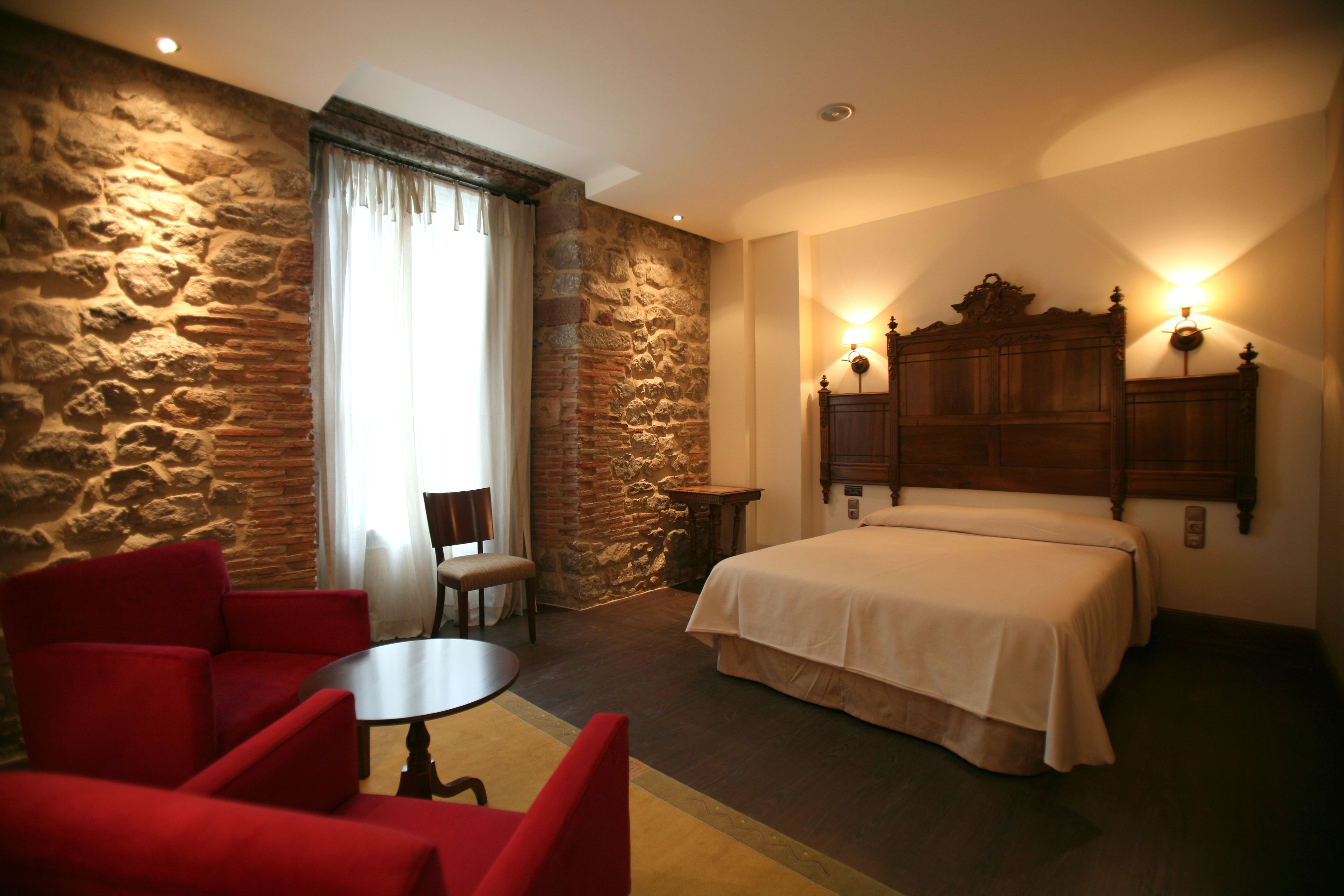 Habitación de hotel con pared de piedra, una cama con cabecera de madera, un sillón rojo, una mesita y una silla.
