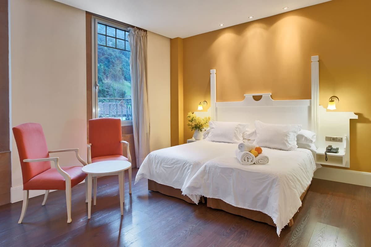 Une chambre d’hôtel avec un lit, une chaise, une petite table, une table de chevet avec une lampe et une fenêtre avec des rideaux.
