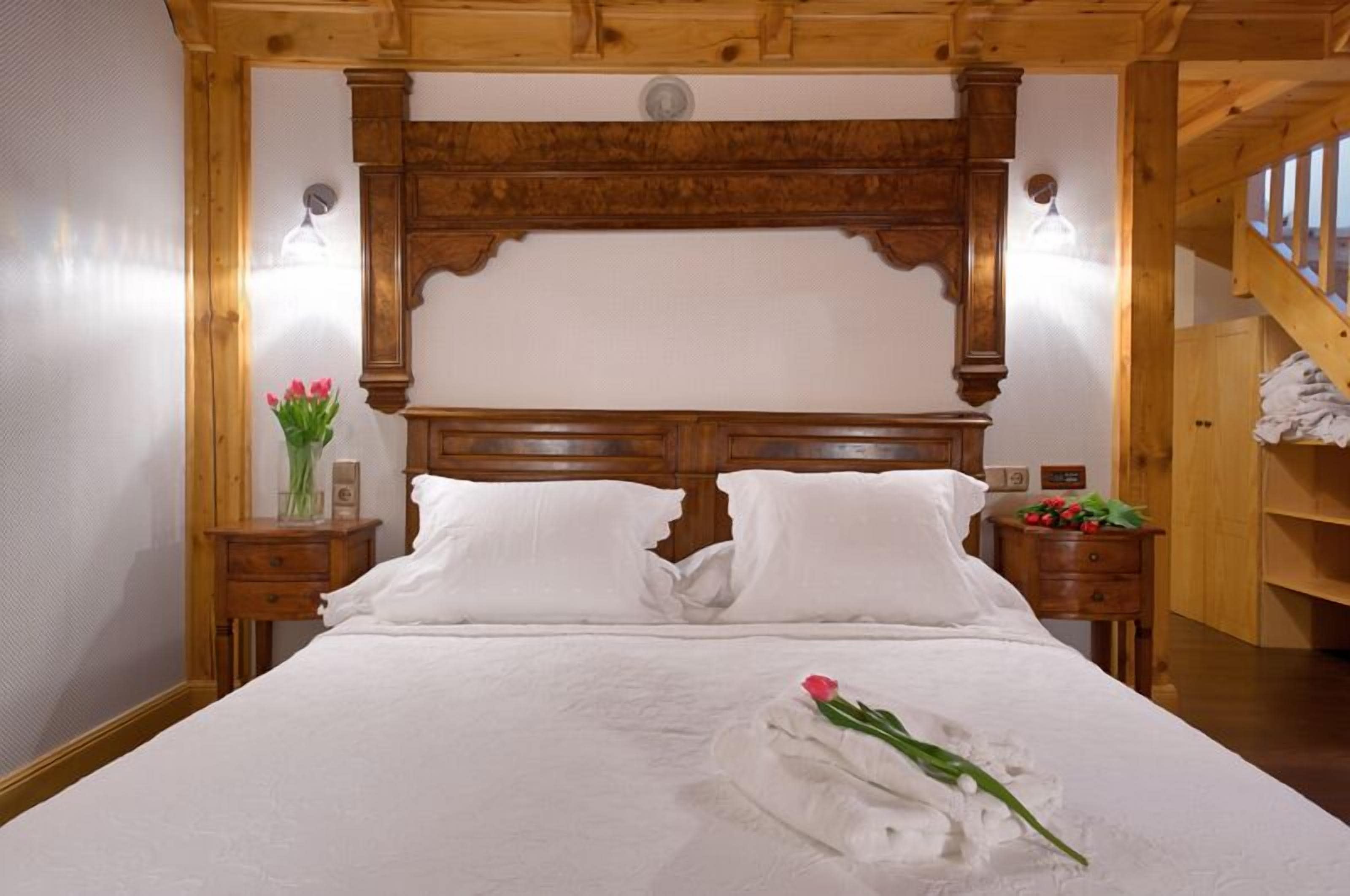 Ein Hotelzimmer mit Holz-Kopfteil, zwei weißen Kissen, einem ordentlich bezogenen Bett, einem Nachttisch mit Blumen und einer Holztreppe im Hintergrund.