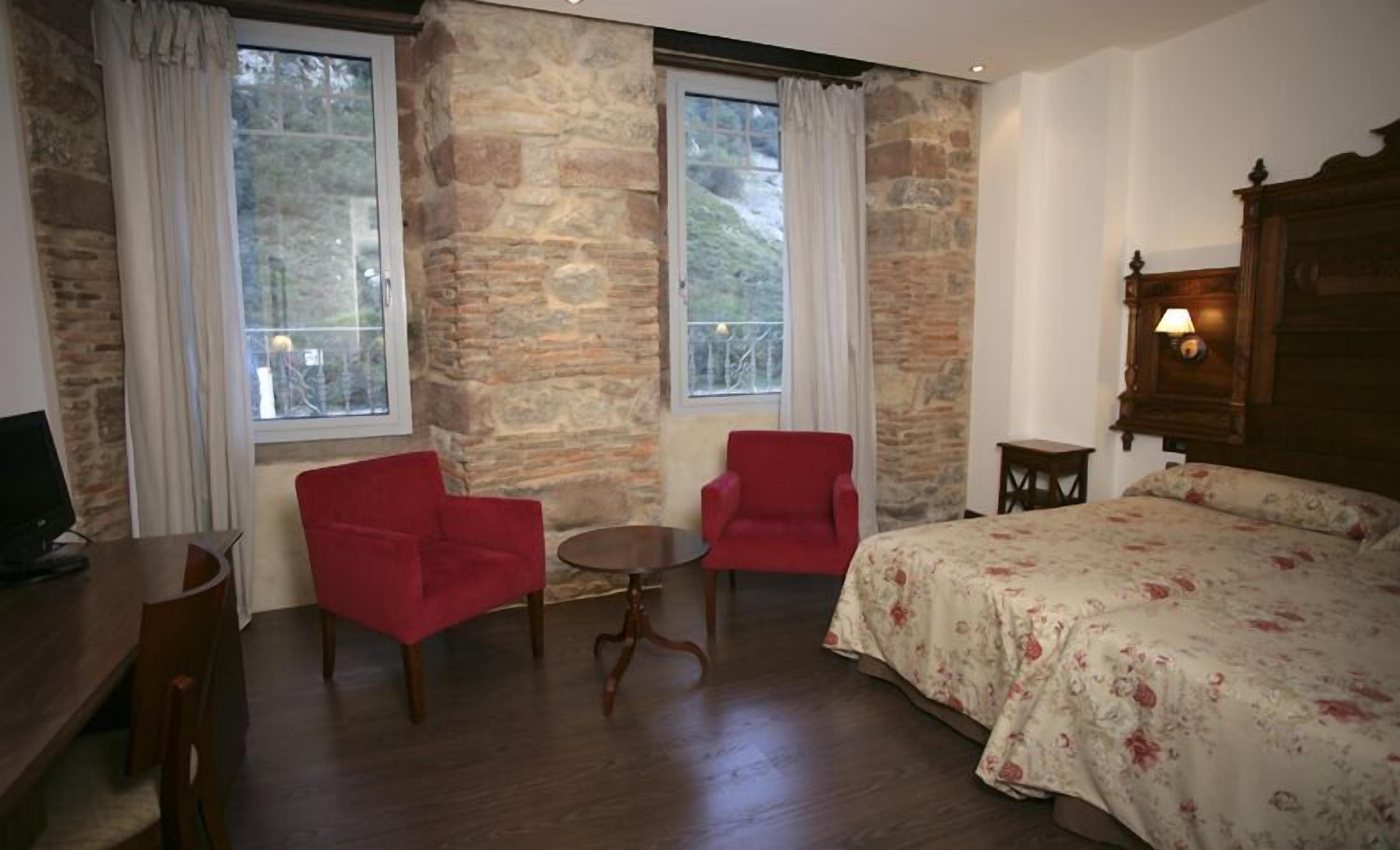 Una habitación con una cama, dos sillas rojas, una mesita, un escritorio y una lámpara.